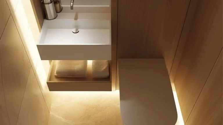 A designer tiny bathroom featuring a wall-mounted sink and faucet, a lit shelf with folded towels underneath, and a toilet adjacent, all adorned with wood paneling and ambient lighting—a perfect space to spend money on thoughtful luxury.