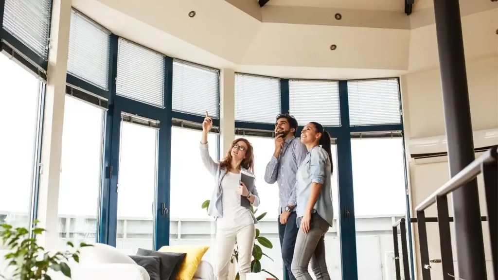 Three people are standing in a spacious room with large windows, and one person, a real estate developer, is pointing upwards.