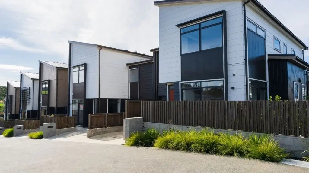 Row of modern, two-story houses with large windows, a wooden fence in front, and a neatly landscaped area with greenery. Built by a legitimate real estate developer, the homes reflect quality craftsmanship under clear skies.
