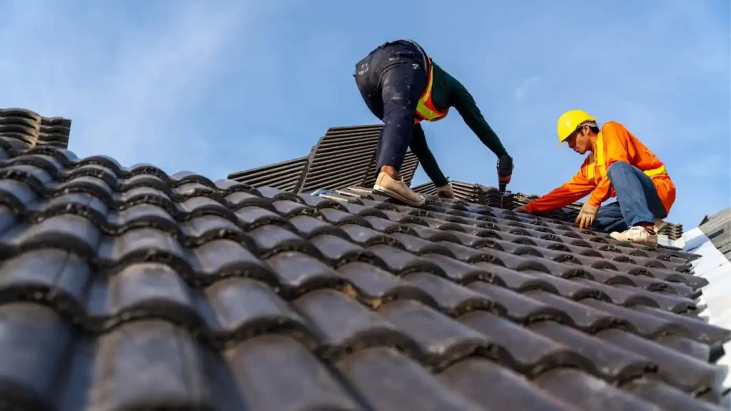 Two construction workers in safety gear install roof tiles on a building under a clear blue sky, showcasing the importance of roofing safety provided by professional roofing services.