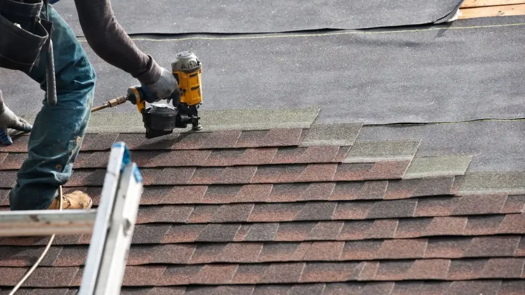 Close-up of a person using a yellow nail gun to safely install shingles on a roof. A ladder is visible in the foreground, emphasizing the need for Professional Roofing Services to ensure quality and safety.