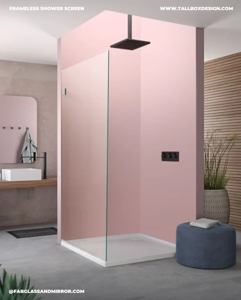 Modern bathroom with a fixed shower screen, large rectangular showerhead, pink accent wall, wooden vanity with a white sink, mirror, and a blue pouf. Text on the image includes website URLs.
