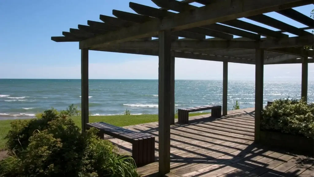 A wooden pergola with benches overlooks a sunny coastal scene with clear blue skies and calm ocean waters, embodying the essence of New Era living.
