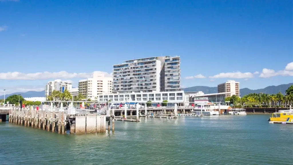 A waterfront with docks, a modern multi-story building, several other buildings, and boats under a clear blue sky highlights the prime real estate of these waterfront residences.