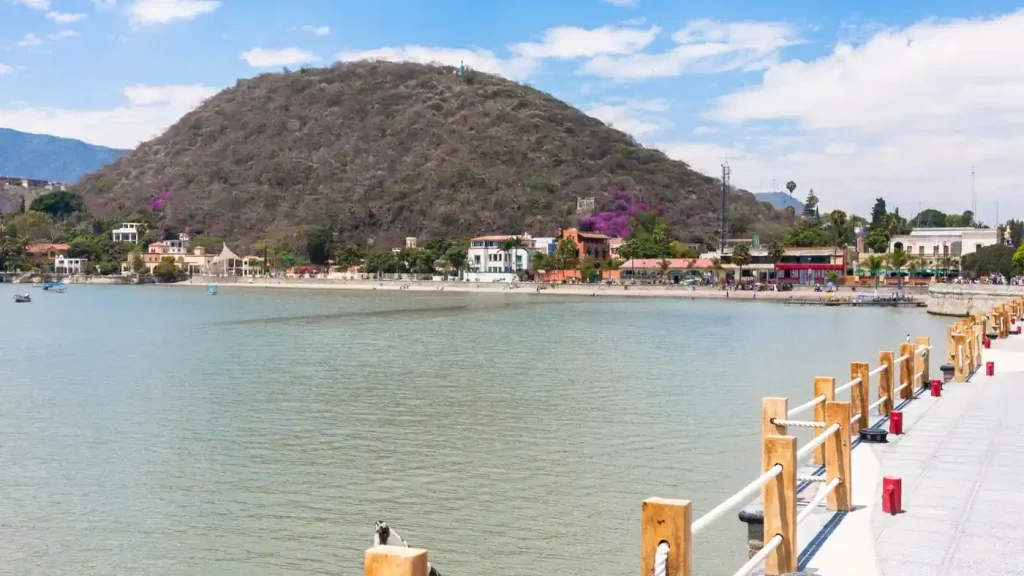 A scenic view of a lake with a wooden dock in the foreground and a small town and hill covered in vegetation in the background under a partly cloudy sky offers an idyllic setting for those buying waterfront property.