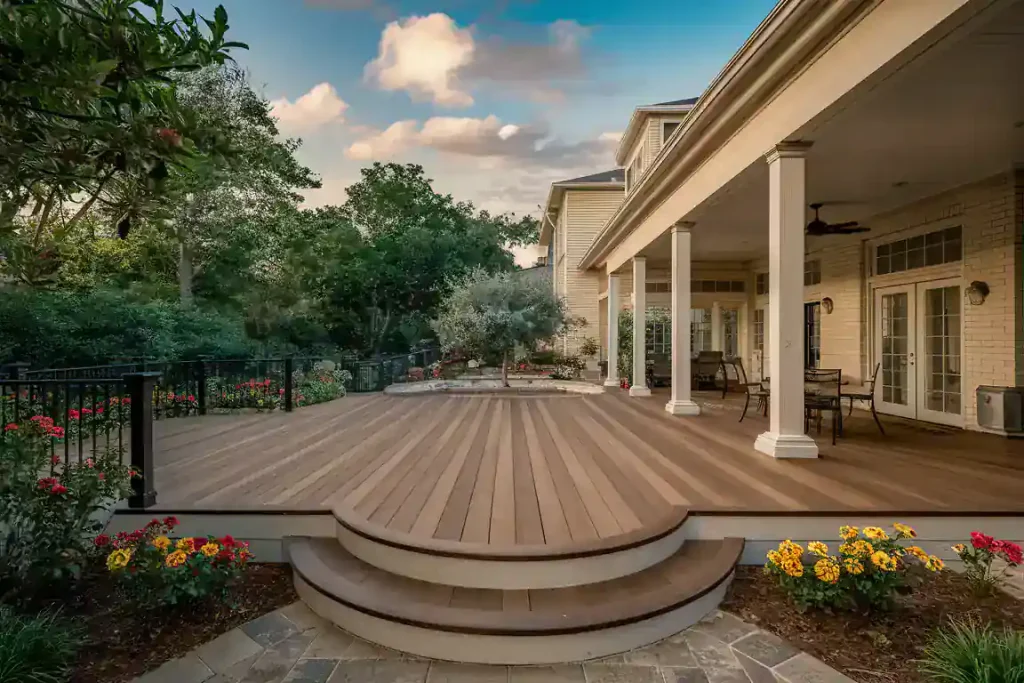 Large patio deck with round steps, covered section, outdoor furniture, and a garden with trees and colorful flowers—a perfect retreat while tackling the challenges of selling an old house.