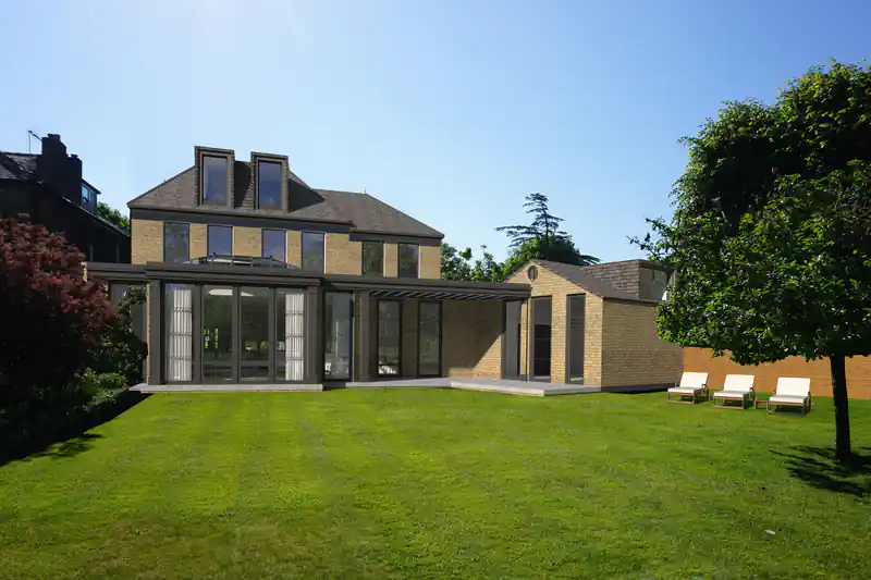 A modern two-story house with a large glass extension and a well-maintained lawn, the result of meticulous pre-construction planning, featuring three white lounge chairs on the right side.