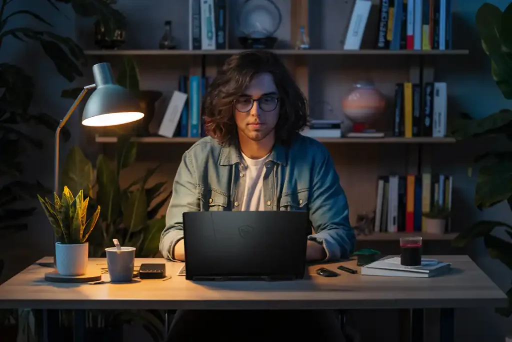 A person with long hair and glasses sits at a desk, immersed in the pre-construction planning process on a laptop. The desk is illuminated by a lamp and adorned with books, plants, a phone, and a cup. A bookshelf stands in the background.