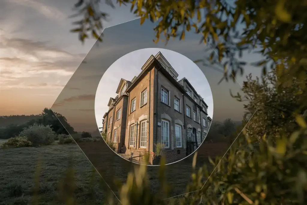 A large, old brick building stands in the countryside, seen through a circular frame with trees around it at sunrise.