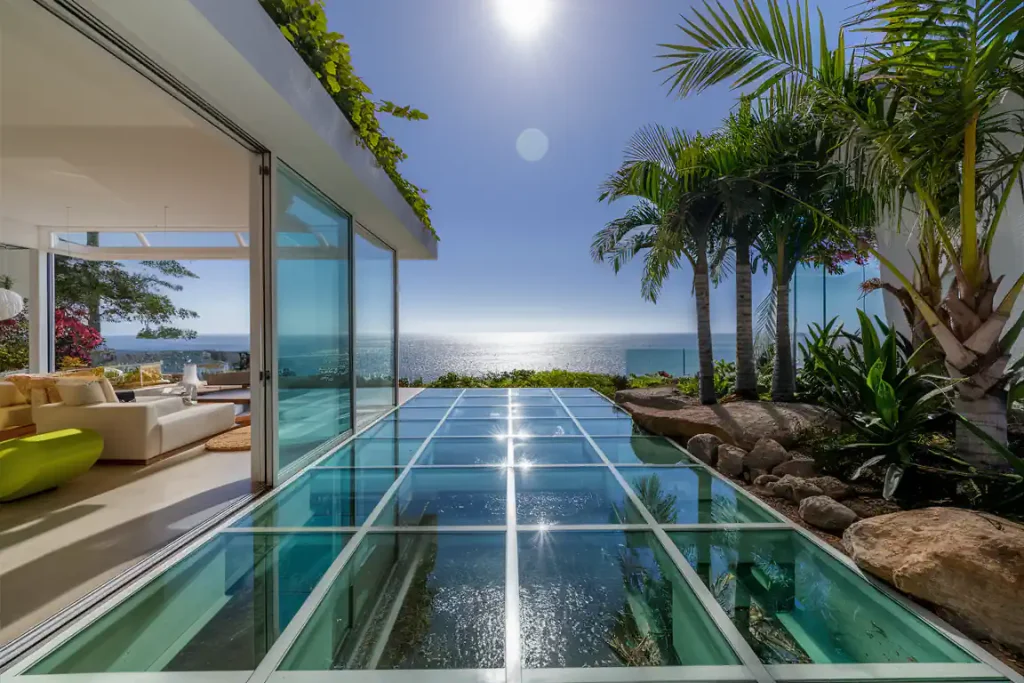 Modern home with glass-floor patio, ocean view, lush plants, and palm trees under a clear blue sky and bright sun. Experience the seamless fusion of nature and design in this meticulously planned haven, born from careful pre-construction planning processes.