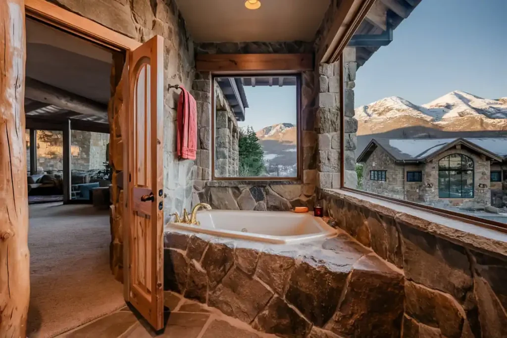 Rustic bathroom with stone walls, wooden door, and a bathtub offering a stunning mountain view through large windows, perfectly crafted.