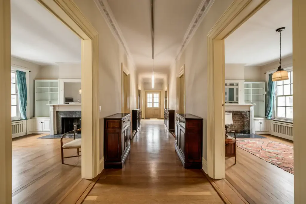 Spacious hallway with wooden flooring, adjacent rooms with large windows, bookshelves, and a brick fireplace—meticulously designed during the pre-construction planning phase.