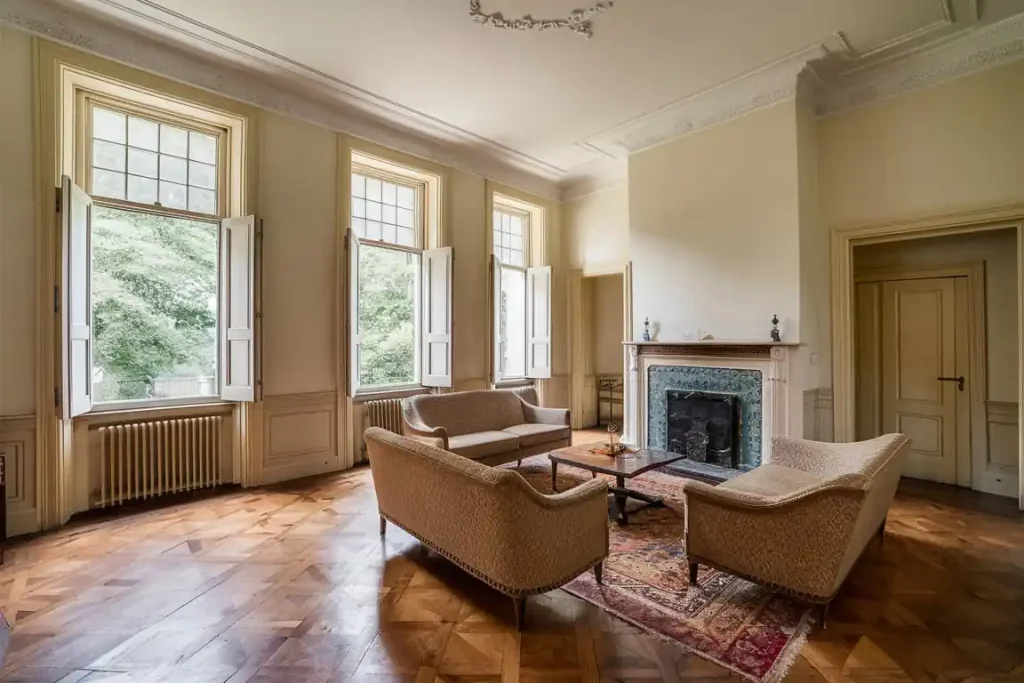 A spacious living room with three windows, classic furniture, a fireplace, and a wooden parquet floor reflects the meticulous attention to detail from the pre-construction planning phase.