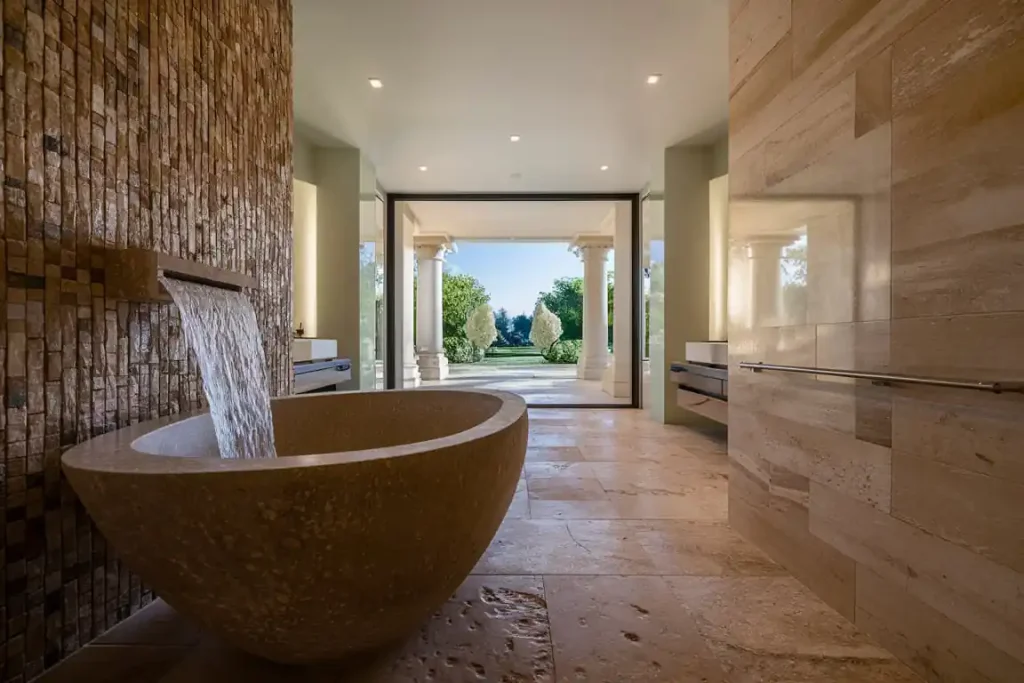 Luxurious bathroom with a stone bathtub and open view of a garden through large glass doors surrounded by columns, expertly designed during the pre-construction planning process.