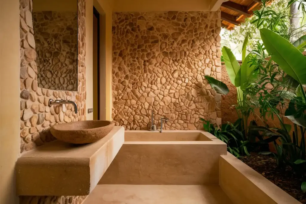 Rustic bathroom with stone walls, a stone sink on a countertop, and a bathtub, surrounded by lush green plants. The seamless design highlights the meticulous