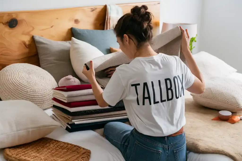 Person in a "TALLBOX" shirt sorting folded fabrics on a bed with pillows, as if meticulously following a planning process, in a cozy room.