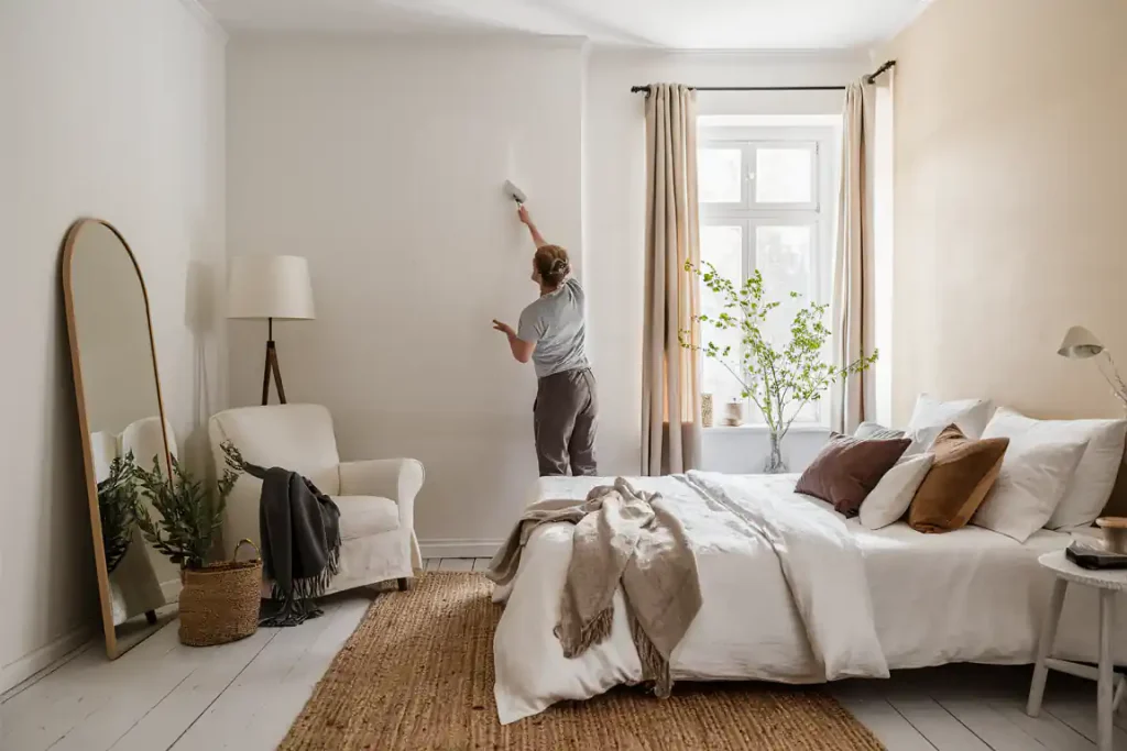 Person cleaning an Organic Modern Bedroom wall with a duster; room with bed, chair, mirror, and plants by the window.