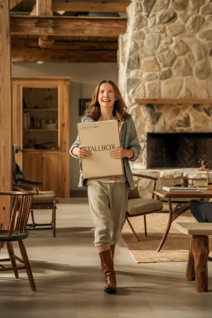 Woman smiling while carrying a TALLBOX in a cozy rustic living room with stone fireplace and wooden furniture, showcasing an organic interior style.