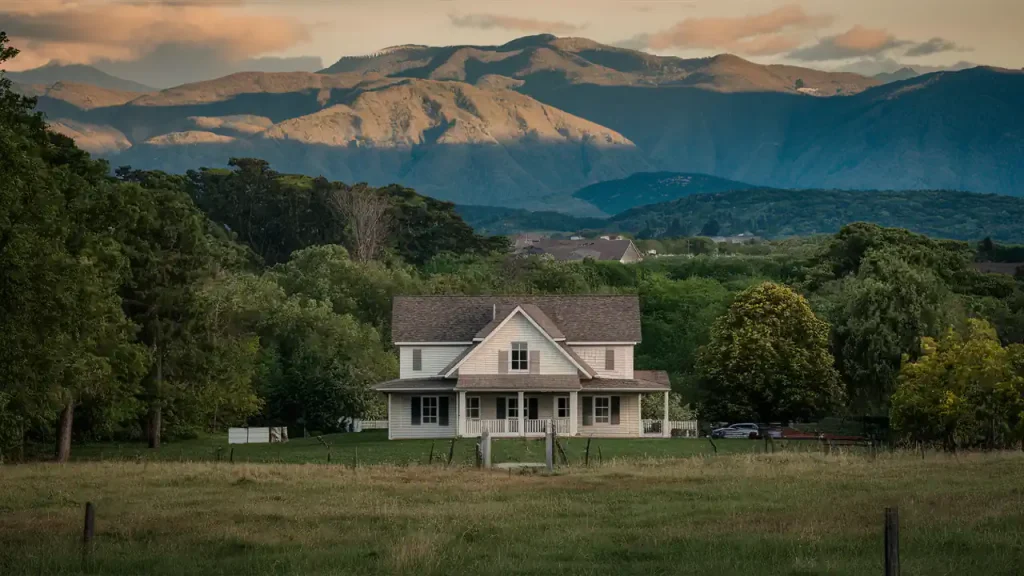 A white house with a porch in a grassy field, set against a backdrop of large, sunlit mountains and a partly cloudy sky—an idyllic scene that reminds you just how rewarding the home buying steps can be.