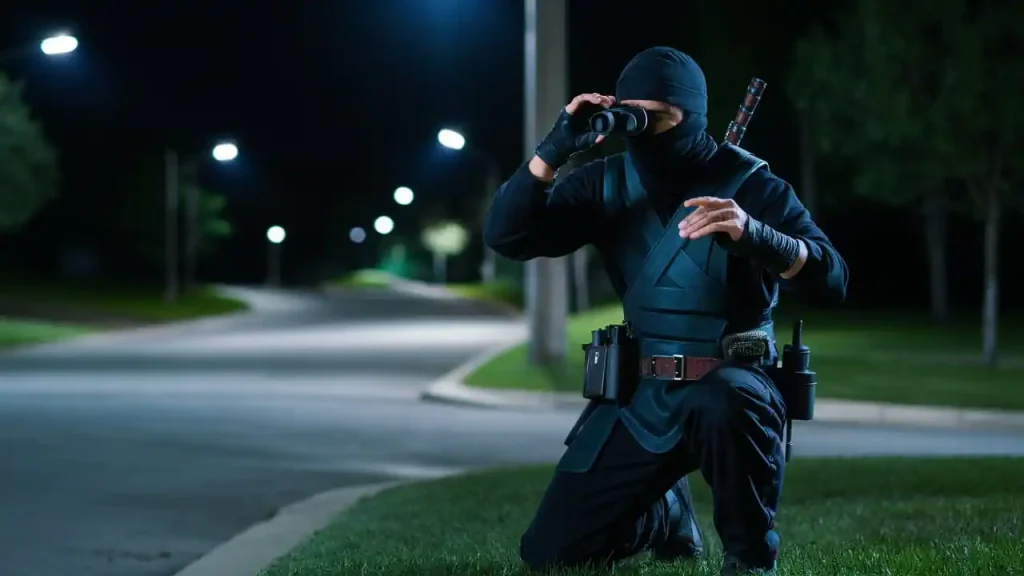 A ninja in black attire kneels on grass at night, looking through binoculars on a deserted street with streetlights, much like a cautious buyer scrutinizing home buying steps.
