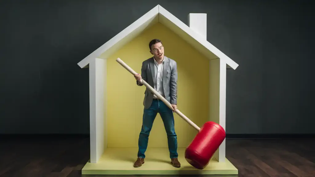 A man in a suit jacket and jeans holds a giant red mallet while standing inside a house-shaped structure, perhaps contemplating the buying a home checklist.