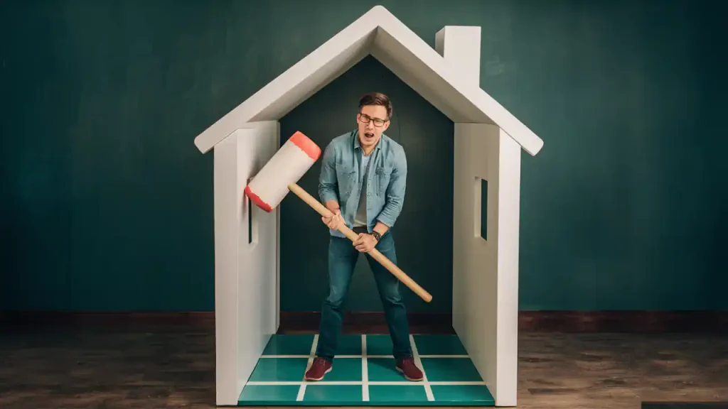 A man in a denim outfit holding a large mallet is standing inside a small model house with green walls and a checkered floor, perhaps symbolizing the first steps to purchasing a home.