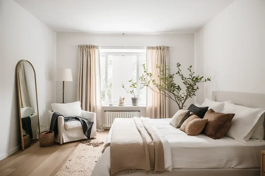 A cozy, Organic Modern Bedroom with a bed, pillows, a chair, mirror, plants, and a large window with beige curtains. The neutral color scheme enhances the organic design, creating a serene and inviting atmosphere.