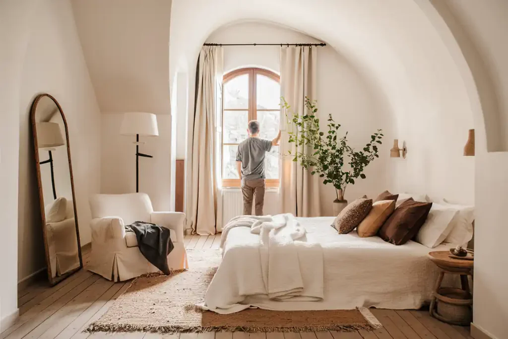A man stands by a large window in a cozy, minimalist bedroom with modern decor, featuring a double bed, armchair, and full-length mirror.