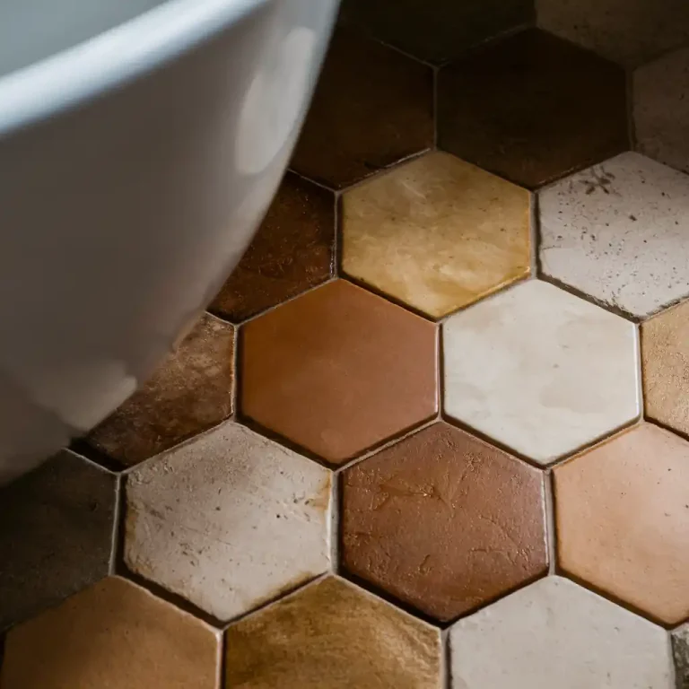 Close-up of a tiled floor with hexagonal tiles in varying shades of brown and beige, creating a warm, organic feel next to a sleek white curved object in a modern bathroom.
