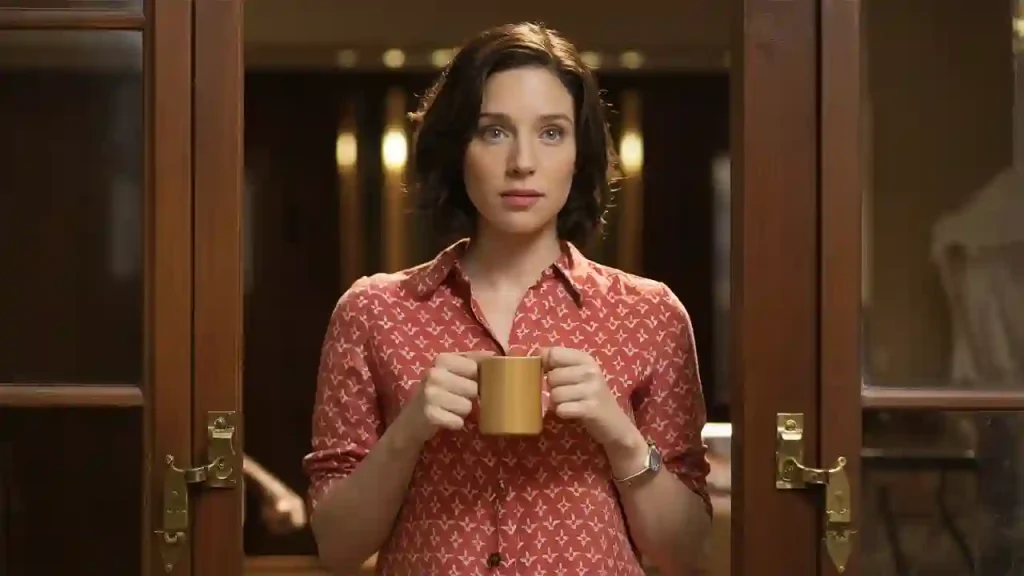 A woman in a red patterned shirt holds a coffee mug, standing in a doorway and looking forward, perhaps considering small fixes to boost her apartment value.