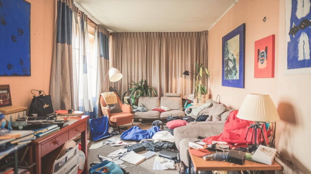 A cluttered living room with scattered clothes and objects, a beige-colored sofa, and a mix of decorations on the walls could benefit from stylish storage solutions.