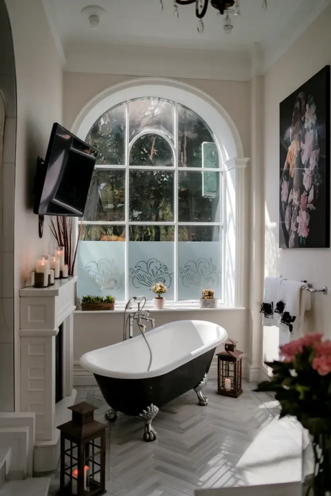 Elegant bathroom with freestanding bathtub, large arched window, candles, wall art, and towel rack. The chic bathroom flooring adds a stylish touch while combining various flooring materials to highlight the room's aesthetic charm.