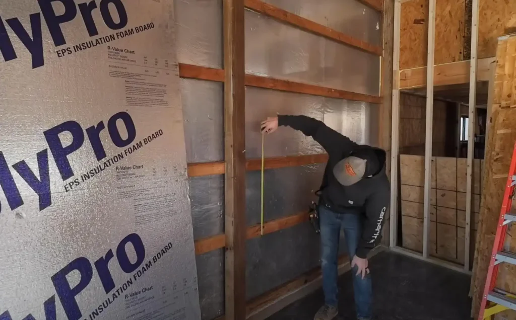 A person is meticulously measuring a wooden wall frame inside a bustling post-frame building construction site, with insulating materials clearly visible in the background.