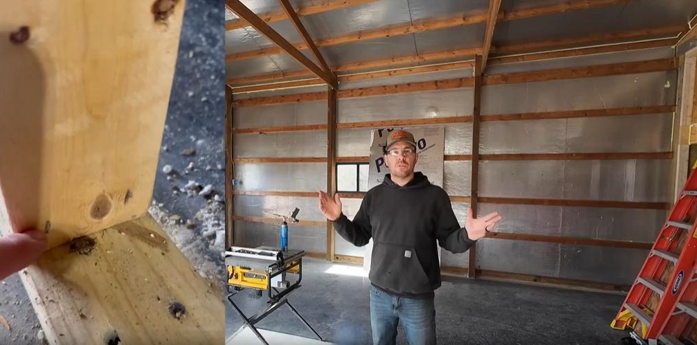 A person in a partially constructed wooden shed, wearing a black hoodie, stands amidst tools and a ladder, preparing for the next phase of the pole barn's development.
