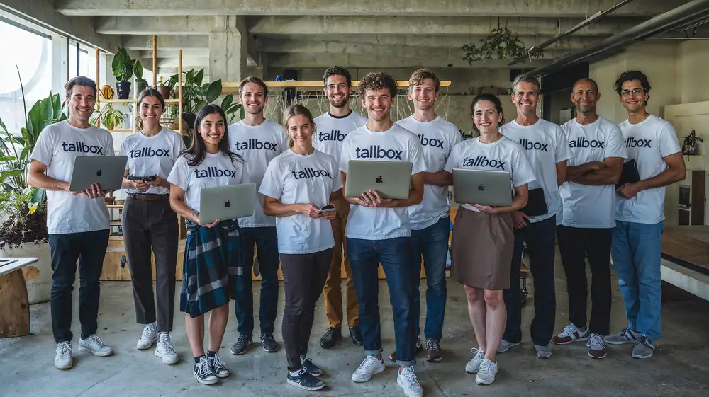 A diverse group of 11 people in matching "tallbox" shirts, each holding a student laptop, smiles in an office, ready to tackle architecture competitions together.