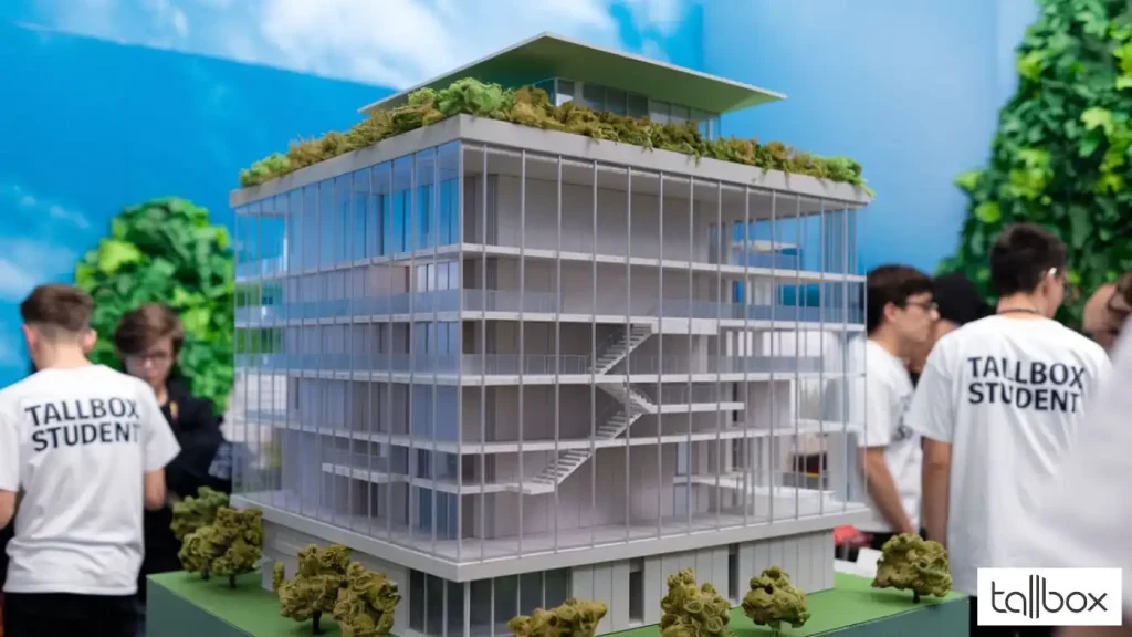 A model of a modern building with trees on each level stands prominently, surrounded by eager students in "Tallbox Student" shirts, perhaps preparing for Architecture Competitions.