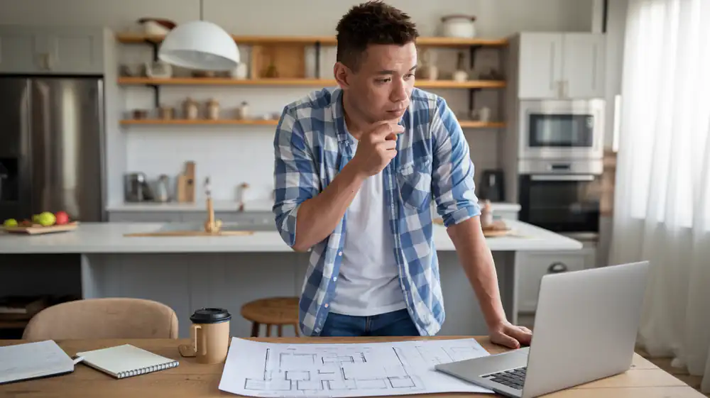 A man stands in his kitchen, thoughtfully gazing at a laptop with blueprints sprawled on the table. He's considering a Reno project and exploring Home Improvement Loans to bring his vision to life.