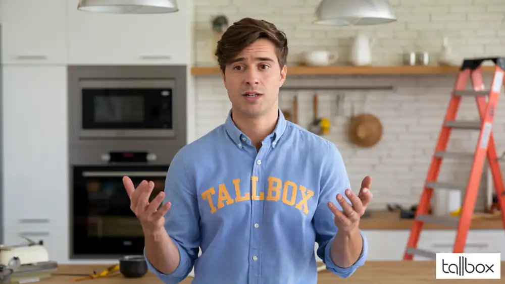 A man in a blue "TALLBOX" shirt gestures in a modern kitchen, alongside a red ladder, showcasing the expertise of remodel contractors.