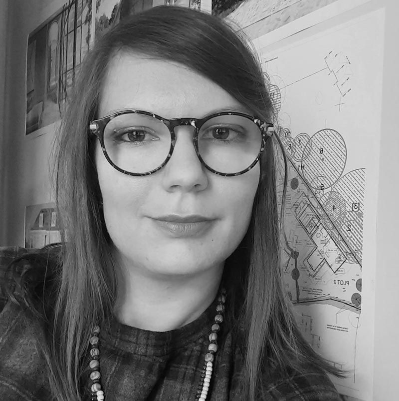 A woman in a plaid outfit and beaded necklace, stands confidently in front of architectural drawings. Her glasses reflect her focus on virtual staging intricacies, even captured in this timeless black and white photo.