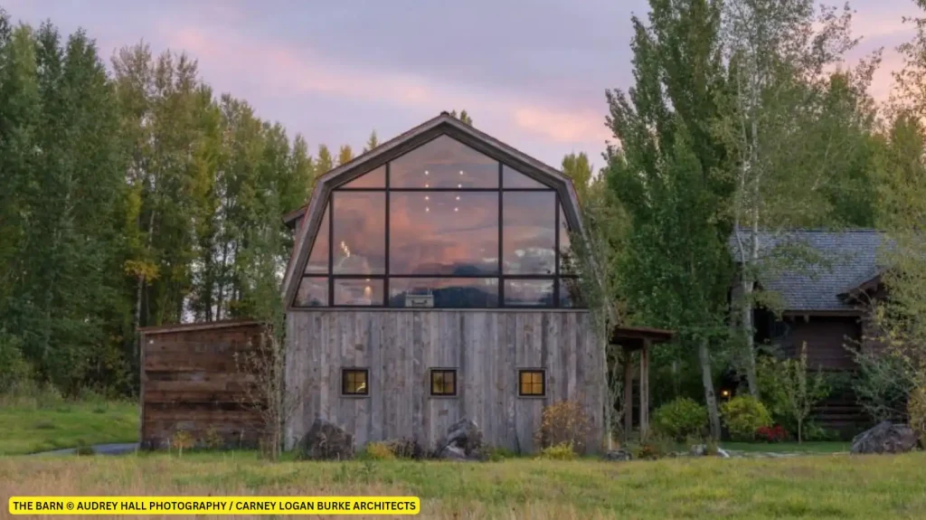 A stunning example of barnhouse architecture, this rustic barn features a large glass front, nestled amidst trees and grass under a colorful evening sky.