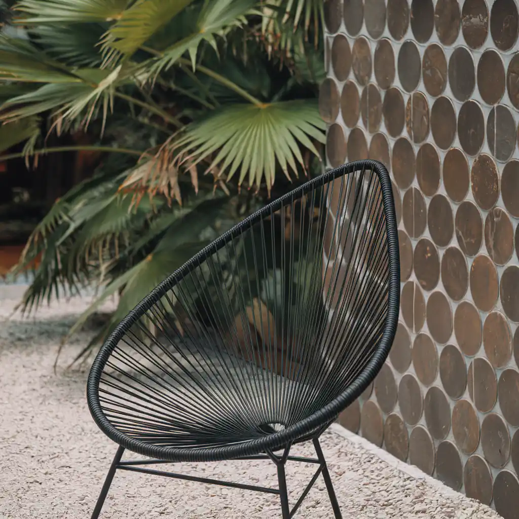 Modern black wire chair on gravel patio, with a textured wall and palm leaves in the background, provides a serene spot for contemplating how to choose swimming pool tiles that complement your outdoor oasis.