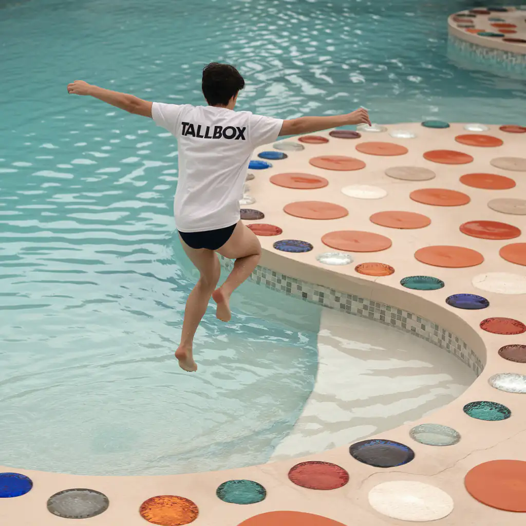 A person wearing a TALLBOX shirt leaps into a pool adorned with an exquisite selection of colorful circular tiles.