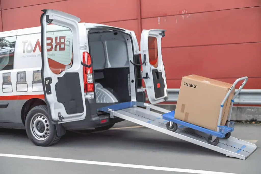 Van with a ramp unloading a large box of packing essentials on a blue cart outside a red building.