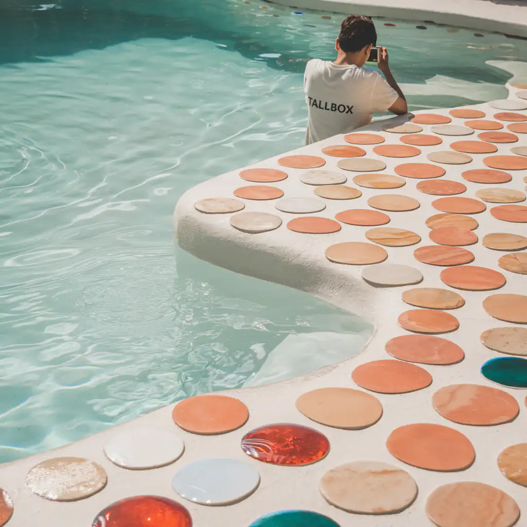 A person sits at the edge of a vibrant swimming pool, capturing the moment with their smartphone. The colorful array of swimming pool tiles adds a stunning backdrop to the scene, showcasing an exquisite swimming pool tile selection.