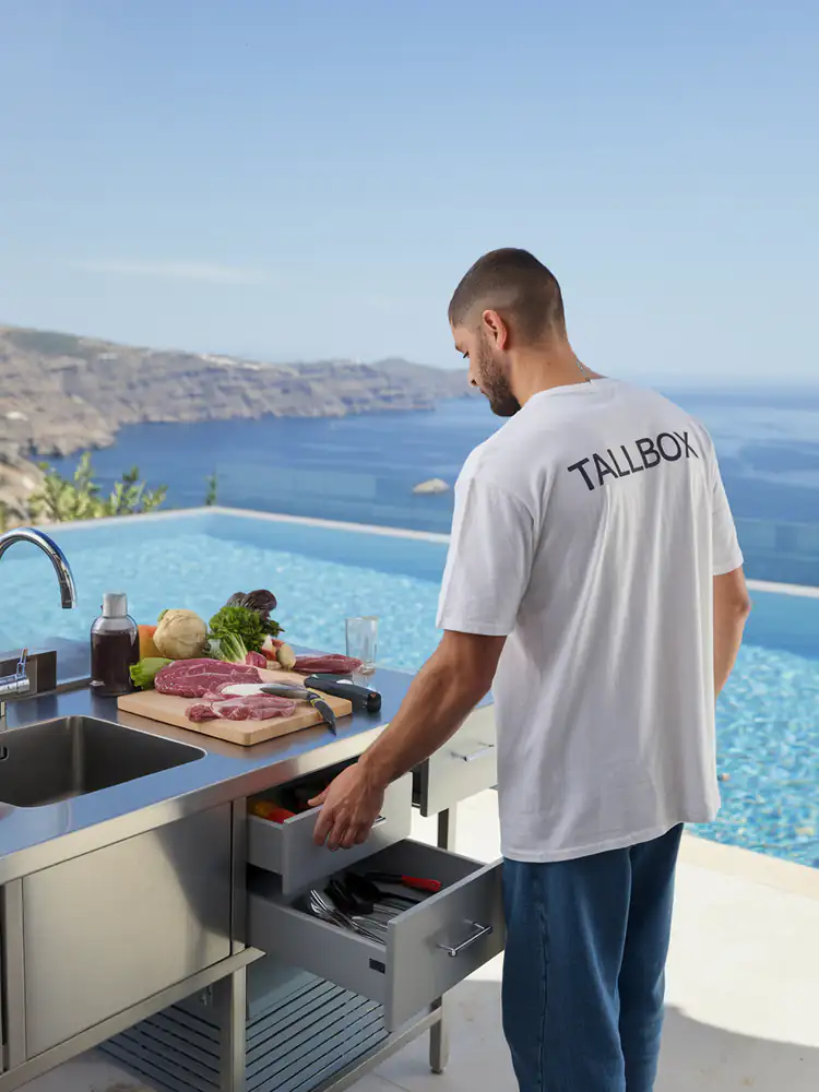 Man cooking in a beautifully designed outdoor kitchen by a pool, with a stunning ocean view in the background.