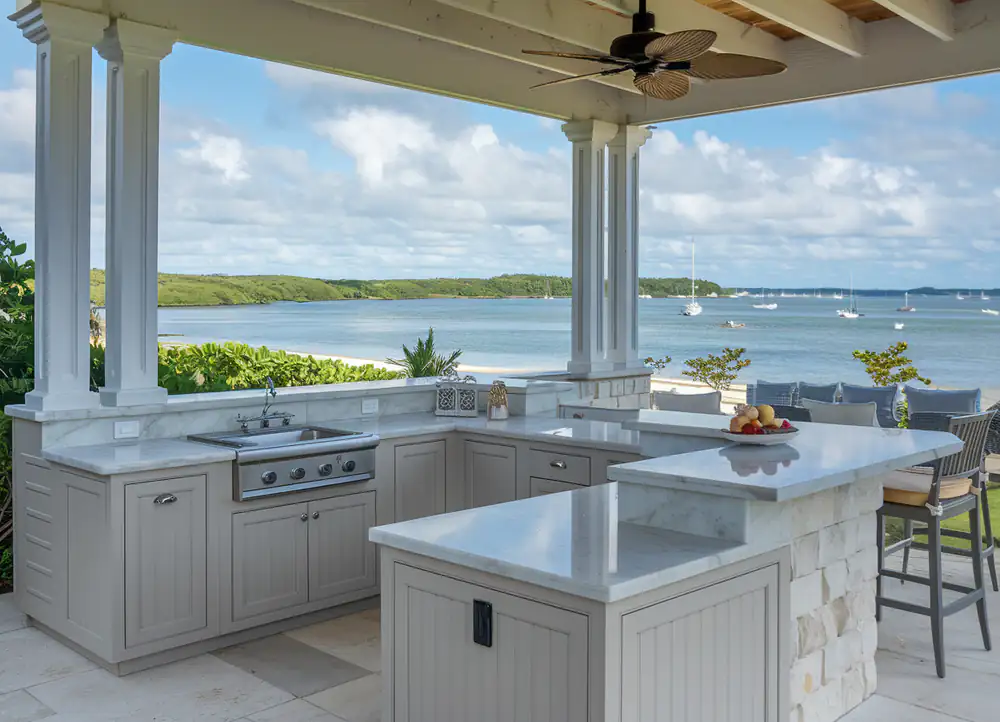Design your dream outdoor kitchen with beige cabinets, marble countertops, and a stunning lake view. A ceiling fan gently hums above while tables are elegantly set with fruit bowls.