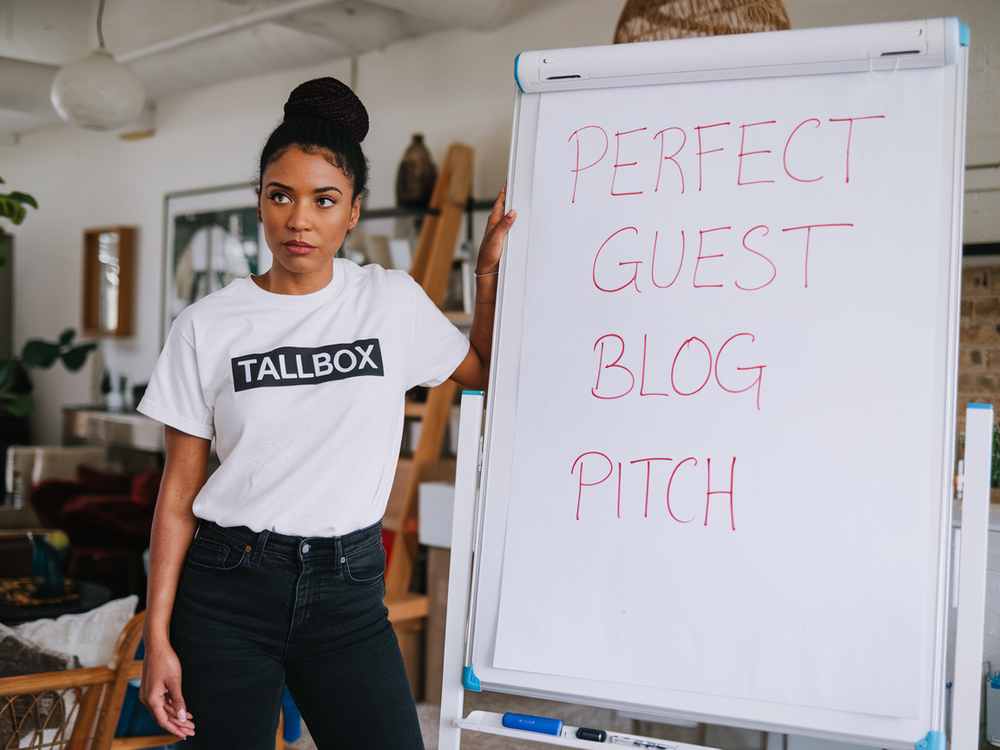 An architect in a "Tallbox" shirt stands beside a flip chart with "PERFECT GUEST BLOG PITCH" written on it, skillfully blending design insight with guest blogging strategies.