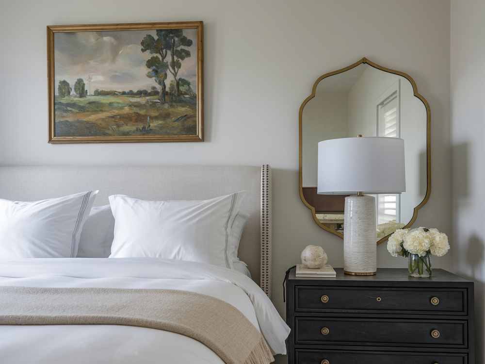 Experience the tranquility of a guest bedroom featuring a plush bed adorned with crisp white linens. A soft lamp casts a warm glow on flowers resting atop a dark nightstand, while an elegant mirror and serene landscape painting complete this luxurious and inviting space.