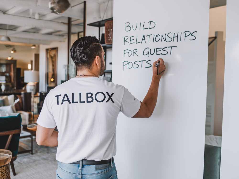 Wearing a shirt labeled "TALLBOX," a person writes "Build relationships for SEO guest posts" on the whiteboard, emphasizing strategies that could benefit both real estate agents and bloggers in the realm of guest blogging.