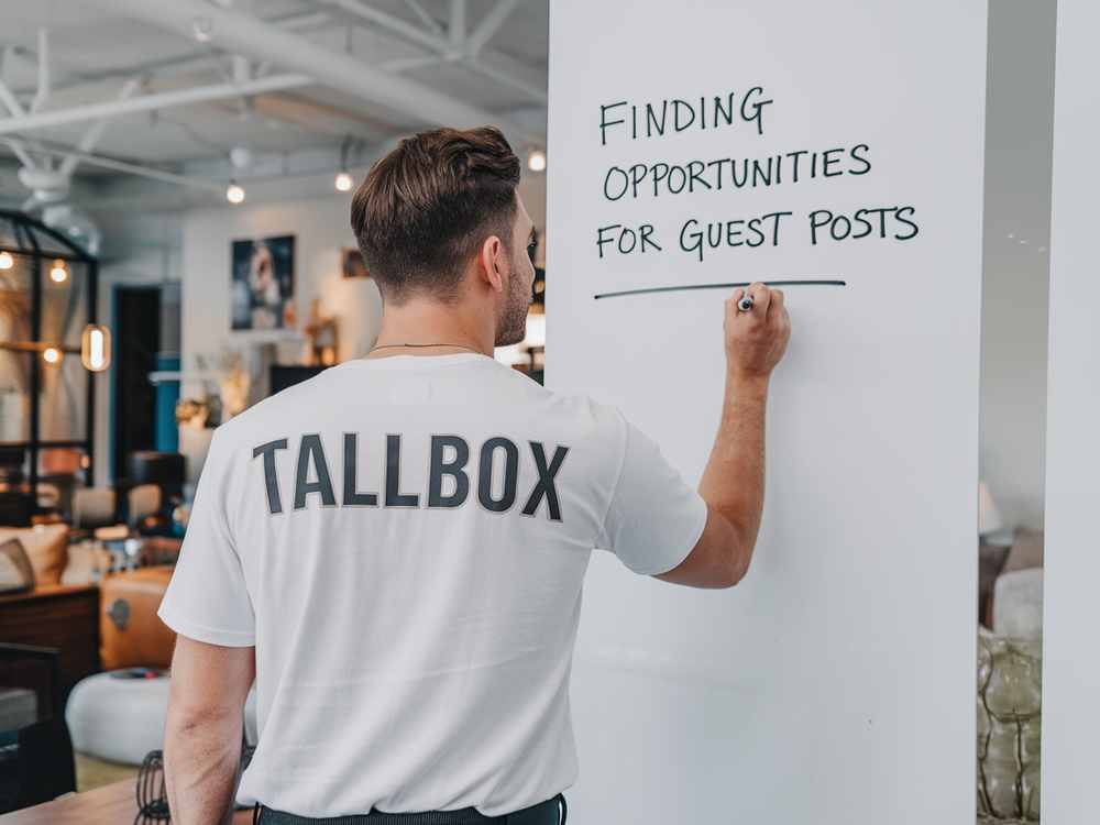 Man in a "Tallbox" shirt writes "Finding opportunities for guest blogging" on a whiteboard in an office, strategizing the perfect approach to enhance visibility through SEO techniques.